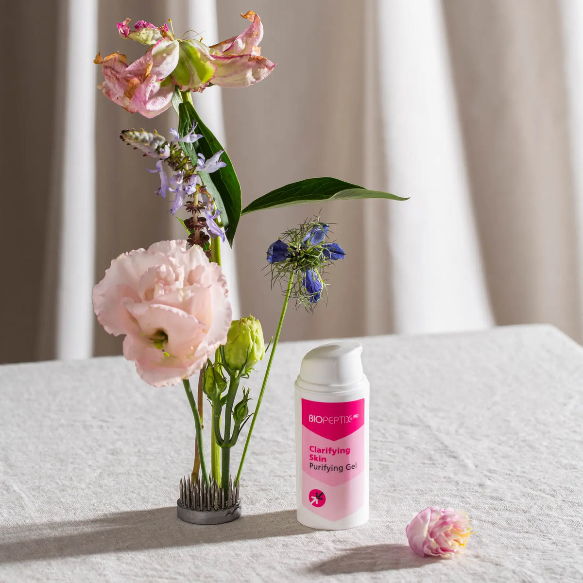 סרום לטיהור ואיזון העור Clarifying Skin של Biopeptix, לצד אגרטל פרחים ורודים וכחולים על משטח פשתן